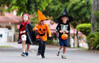 Three children in Halloween costumes, including a witch, a pumpkin, and a vampire, walk outside holding candy buckets during the daytime.