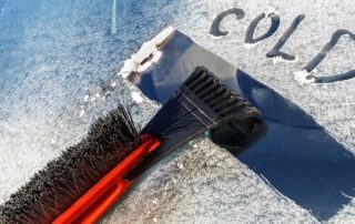 A close-up of a car windshield being scraped free of frost, with the word COLD written in the frost and an ice scraper and brush in use.