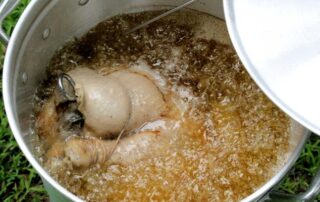 Two chicken legs are being deep-fried in hot oil inside a large metal pot, partially covered by a lid.
