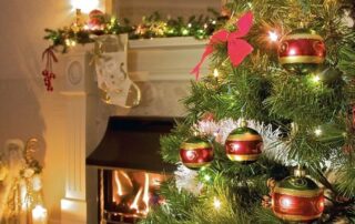 A decorated Christmas tree with red and gold ornaments stands near a lit fireplace and a festive mantel adorned with stockings and garland.