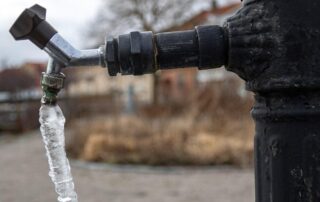 A black outdoor water faucet with a long icicle hanging from the spout, indicating freezing temperatures.