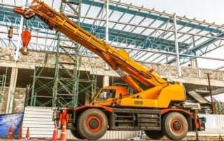 A yellow mobile crane is parked at a construction site in front of a partially built structure with steel framework and scaffolding.