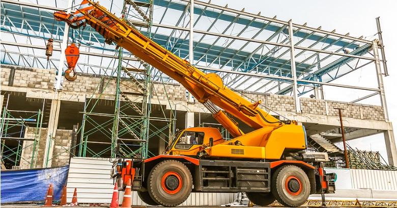 A yellow mobile crane is parked at a construction site in front of a partially built structure with steel framework and scaffolding.