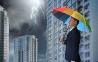 A man in a suit holds a rainbow-colored umbrella amid tall buildings as lightning strikes in the cloudy sky nearby.