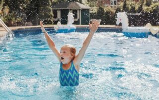 A young girl in a blue swimsuit raises her arms and splashes water while playing in an outdoor swimming pool.