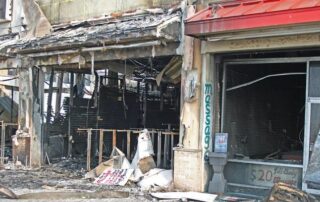 The image shows the burnt and heavily damaged storefronts of businesses, with debris scattered on the ground and exposed building interiors.