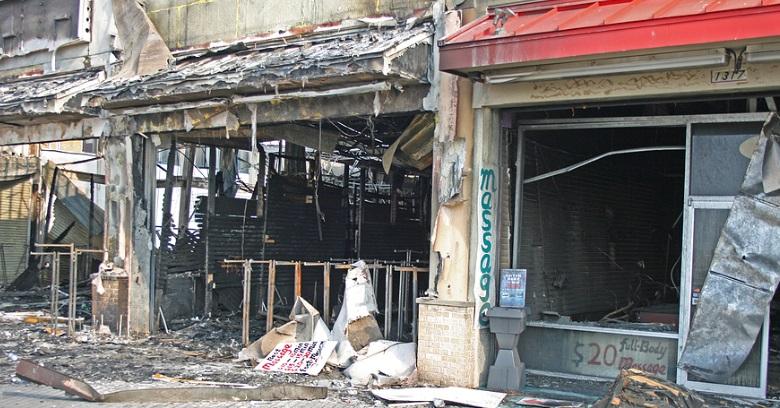 The image shows the burnt and heavily damaged storefronts of businesses, with debris scattered on the ground and exposed building interiors.