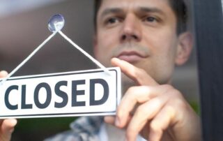 A person holds up a sign that reads CLOSED against a window, indicating that the establishment is not open.