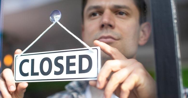 A person holds up a sign that reads CLOSED against a window, indicating that the establishment is not open.