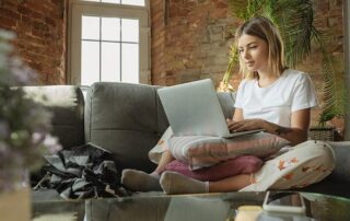 A woman sits cross-legged on a sofa using a laptop, with a pillow on her lap and a window behind her, in a room with exposed brick walls and plants.