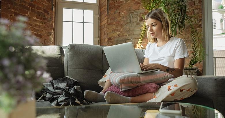 A woman sits cross-legged on a sofa using a laptop, with a pillow on her lap and a window behind her, in a room with exposed brick walls and plants.