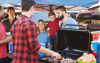A group of people gather around a grill at an outdoor event, with one person cooking burgers and others holding drinks under a canopy.
