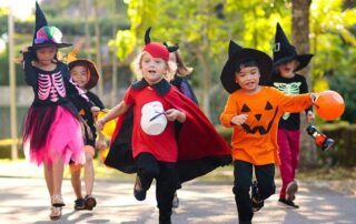 A group of children in Halloween costumes walk and run outdoors during the day, holding candy buckets.