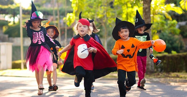 A group of children in Halloween costumes walk and run outdoors during the day, holding candy buckets.
