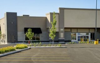 A modern, empty commercial building with black awnings and large windows, surrounded by a paved parking lot and young landscaping.