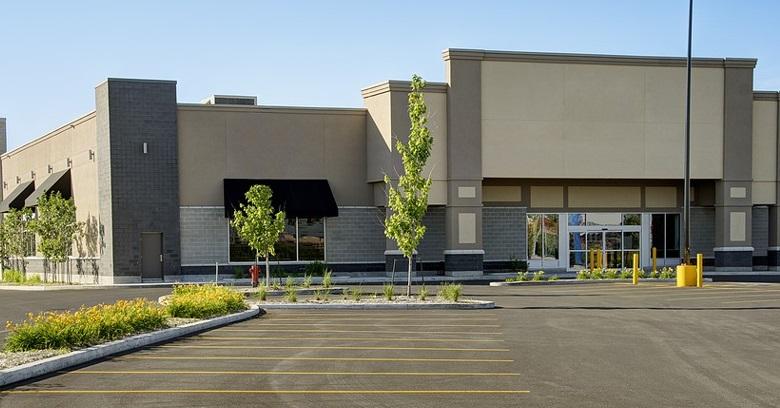 A modern, empty commercial building with black awnings and large windows, surrounded by a paved parking lot and young landscaping.