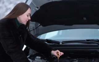A woman wearing a dark coat checks the oil dipstick under the hood of a car.