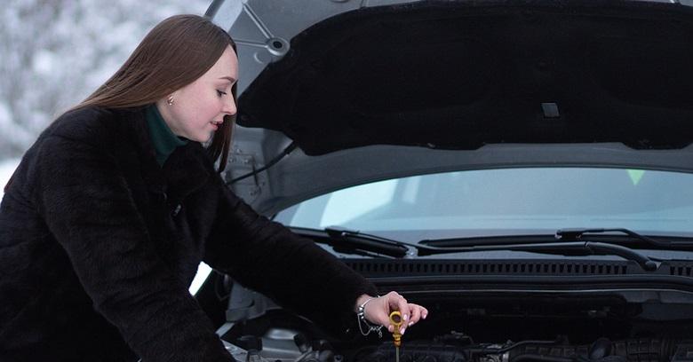 A woman wearing a dark coat checks the oil dipstick under the hood of a car.