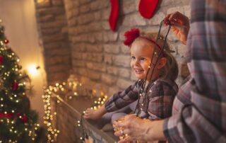 A young girl smiles while sitting on a mantel as an adult beside her holds a string of holiday lights, with a decorated Christmas tree and stockings in the background.