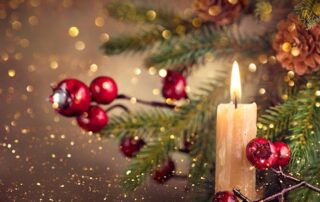 A lit candle sits among pine branches, pinecones, and red berries, with golden bokeh lights in the background.
