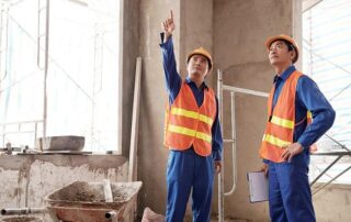 Two construction workers in safety vests and helmets stand inside a building under construction; one points upward while the other holds a clipboard.