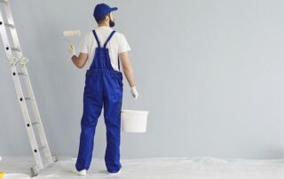 A person in blue overalls holds a paint roller and a bucket, standing near a ladder in front of a light gray wall.