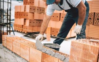 A construction worker in gloves and overalls lays mortar and aligns bricks while building a wall at a construction site.