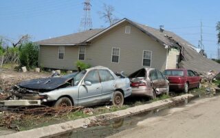 Three damaged cars are lined up in front of a house with a collapsed roof, surrounded by storm debris and muddy ground.