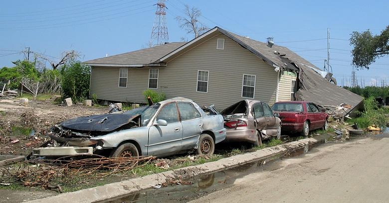 Three damaged cars are lined up in front of a house with a collapsed roof, surrounded by storm debris and muddy ground.