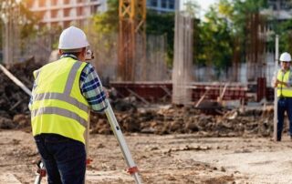 Two construction workers wearing safety vests and helmets use surveying equipment at a building site with rebar columns and dirt.