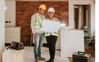 Two construction workers in safety vests and helmets examine blueprints inside a building under renovation, with tools and materials visible around them.