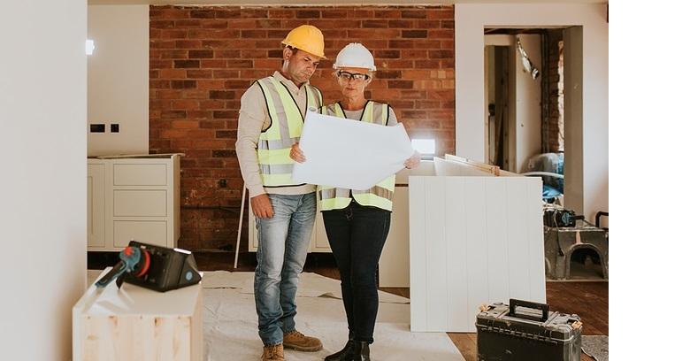 Two construction workers in safety vests and helmets examine blueprints inside a building under renovation, with tools and materials visible around them.