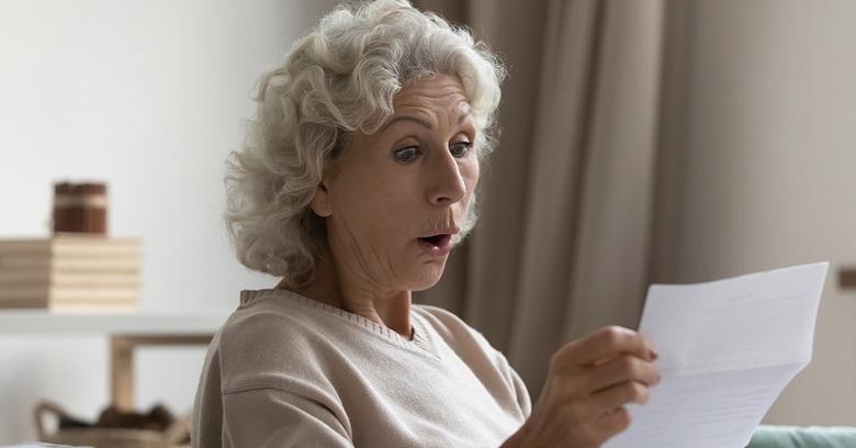 An older woman with curly gray hair looks surprised while reading a piece of paper indoors.