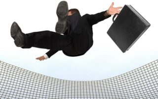 A person in a business suit and holding a briefcase falls backward onto a safety net against a plain background.