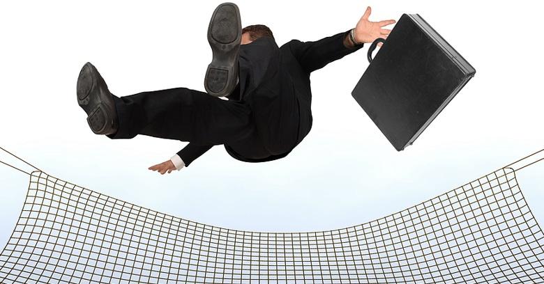 A person in a business suit and holding a briefcase falls backward onto a safety net against a plain background.