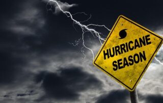 A yellow Hurricane Season warning sign stands against a dark, cloudy sky with a bolt of lightning in the background.
