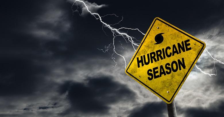 A yellow Hurricane Season warning sign stands against a dark, cloudy sky with a bolt of lightning in the background.
