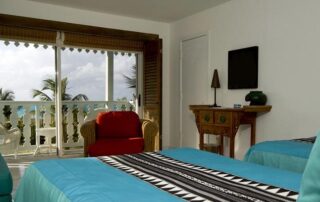 A hotel room with two turquoise beds, a red armchair, TV, small table, and a balcony overlooking palm trees and the ocean.
