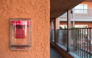 Red fire alarm pull station behind a clear plastic cover mounted on an orange stucco wall in an outdoor apartment corridor.