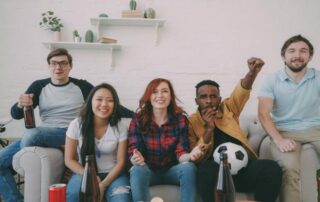Five people sit on a couch watching something intently, with drinks and a soccer ball, suggesting they are watching a sports game.