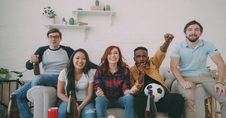 Five people sit on a couch watching something intently, with drinks and a soccer ball, suggesting they are watching a sports game.