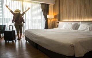 A person stands with arms raised by a window in a hotel room, next to a suitcase, with sunlight streaming through sheer curtains onto a neatly made bed.