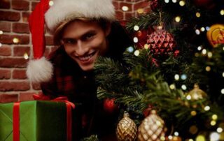 A person wearing a Santa hat smiles while holding a green gift box next to a decorated Christmas tree with gold and red ornaments.