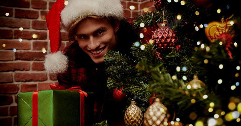 A person wearing a Santa hat smiles while holding a green gift box next to a decorated Christmas tree with gold and red ornaments.