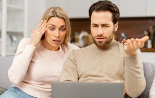 A man and woman sit on a couch looking at a laptop screen, appearing confused and concerned.