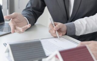 Two people in business attire review and sign a document at a desk, with a laptop and model houses visible in the foreground.