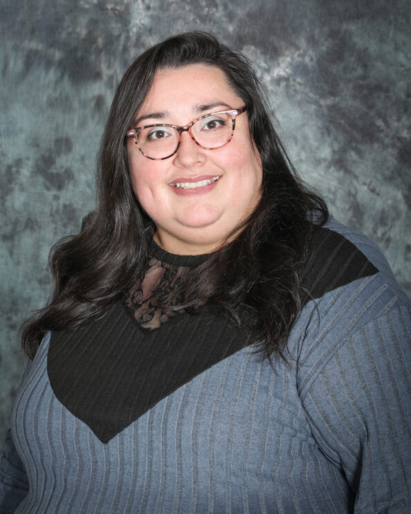 A woman with long dark hair and glasses is smiling, wearing a gray and black top, posed in front of a mottled gray backdrop.