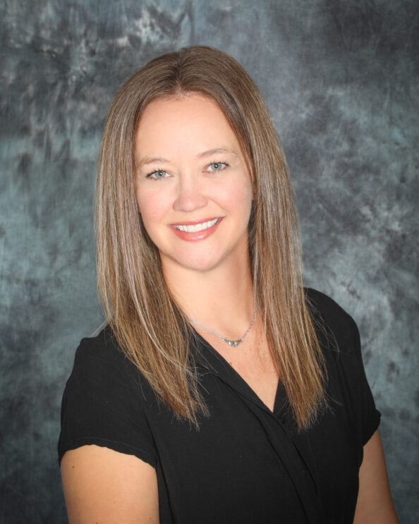 A woman with straight, light brown hair wearing a black top and a silver necklace smiles in front of a mottled gray background.