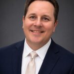 Professional headshot of a man in a navy suit and white tie against a gray background.